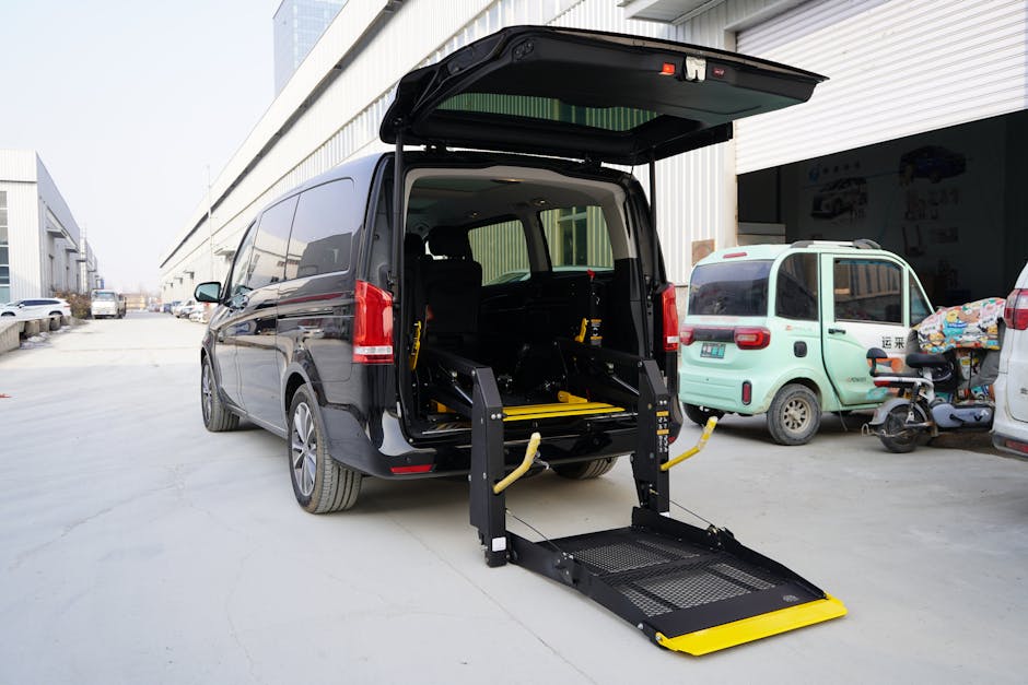A black wheelchair-accessible conversion van with its rear hatch open, equipped with a motorised wheelchair lift platform extending from the ground to inside the vehicle. The lift has yellow safety rails and a mesh surface for secure wheelchair loading. The van is parked on a paved driveway outside a commercial building, with other vehicles including a small light green electric cargo vehicle and a motorcycle nearby. Interior visibility shows space for transporting furniture or household items, suggesting the vehicle is used by Man and Van Kingston for house removals and home relocation services. The environment features an industrial setting with a warehouse-like facade, emphasizing logistics and furniture transport processes associated with packing and moving tasks.
