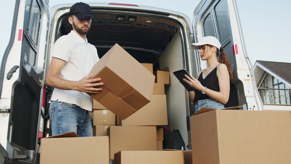 A man and a woman are engaged in a home relocation process involving the loading of cardboard boxes into the back of a white moving van parked outside a residential property. The man, dressed in a white polo shirt, blue jeans, and a dark baseball cap, is holding a medium-sized cardboard box, preparing to place or move it inside the van. The woman, wearing a black sleeveless top, light blue jeans, and a white cap, is standing nearby, focused on a tablet device, possibly coordinating or checking a moving checklist. Inside the van, several cardboard boxes of various sizes are stacked on the floor, some sealed with packing tape, indicating packing and moving preparations. The van is equipped with a dark interior suitable for transporting household items, with a small portion of the driveway or pavement visible at the bottom of the image. In the background, part of a house with a pitched roof and window can be seen under a cloudy sky, highlighting an outdoor residential environment typically associated with house removals, where carefully packing, lifting, and loading processes are underway. Man and Van Kingston professional services might be involved in this furniture transport, ensuring a smooth home relocation transition.