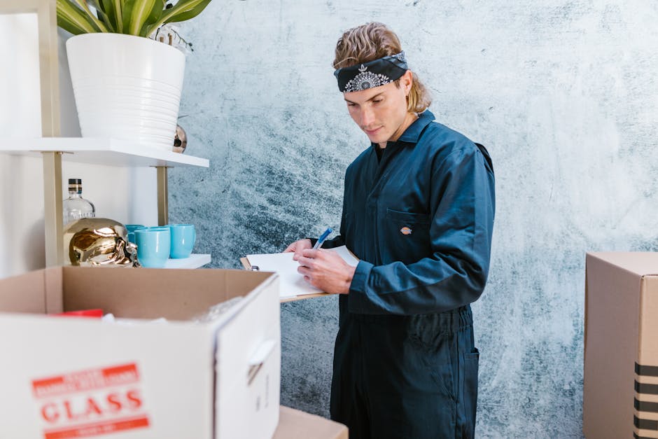 A young man wearing a dark blue work jumpsuit and a black bandana with white patterns is inside a residential or commercial property, using a clipboard and pen to take notes. In front of him, there are open cardboard boxes filled with various packing materials and household items, including a small decorative ceramic object, blue cups, and a clear glass container with a metallic lid. The boxes are positioned on a surface possibly a table or shelf, and the background features a textured, light-colored wall. To the left, there is a white shelving unit holding additional items such as a large white pot with a plant, a clear glass bottle, and some smaller objects. The scene appears to be part of a home relocation or moving process, with visible packing and organization happening during the planning or inventory stage, which is relevant to the house removals services provided by Man and Van Kingston, as indicated in the context of the page about a busy moving day at Bentall Centre in Kingston.