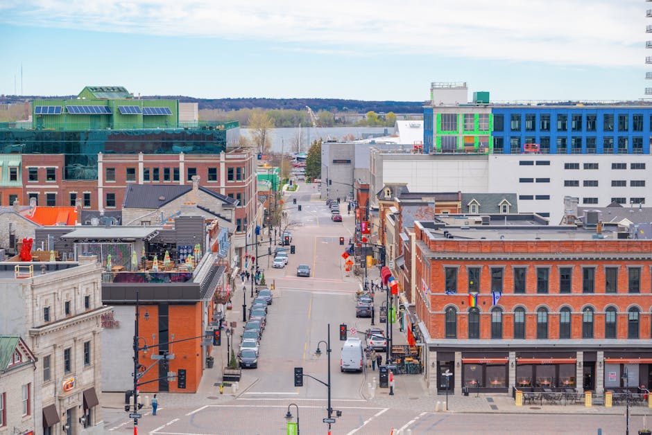 An aerial view of a street in Kingston, showing a mixture of historic and modern buildings with brick facades, retail shops, and commercial offices, some of which feature colorful signage and flags. The street is lined with parked cars and has a few pedestrians walking along the sidewalks. In the background, there are newer multi-storey buildings, including one with a green rooftop structure and large windows, and another with a blue facade. Beyond the buildings, a river with a bridge and distant trees are visible under a partly cloudy sky. This scene captures the urban environment where home relocation and furniture transport activities may take place, often involving moving companies like Man and Van Kingston, who handle packing and loading processes.