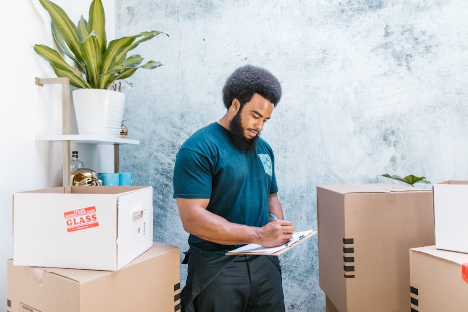 A young man wearing a dark blue work jumpsuit and a black bandana with white patterns is inside a residential or commercial property, using a clipboard and pen to take notes. In front of him, there are open cardboard boxes filled with various packing materials and household items, including a small decorative ceramic object, blue cups, and a clear glass container with a metallic lid. The boxes are positioned on a surface possibly a table or shelf, and the background features a textured, light-colored wall. To the left, there is a white shelving unit holding additional items such as a large white pot with a plant, a clear glass bottle, and some smaller objects. The scene appears to be part of a home relocation or moving process, with visible packing and organization happening during the planning or inventory stage, which is relevant to the house removals services provided by Man and Van Kingston, as indicated in the context of the page about a busy moving day at Bentall Centre in Kingston.