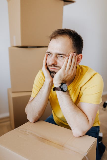 A man with short dark hair and a beard, wearing a yellow T-shirt and a black wristwatch, sits at a table resting his head in his hands with a concerned expression. Behind him, there are several cardboard moving boxes stacked against a white wall, some of which are open, revealing packing paper and plastic wrapping. The setting appears to be an indoor space during a home relocation process, with additional boxes on the floor and a partially visible van or storage area nearby, indicating ongoing furniture transport or packing and moving activities. The lighting is bright and natural, highlighting the materials involved in house removals. This image reflects the stress and logistical challenges associated with last-minute Kingston removals, often managed by companies like Man and Van Kingston.