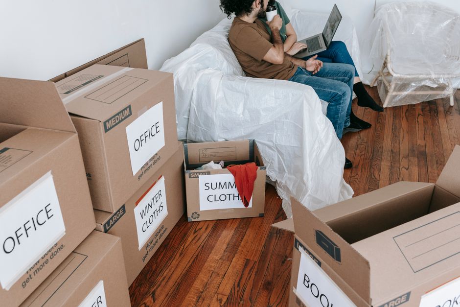 Inside a residential room during the packing stage of a home relocation, several brown cardboard boxes are visible, labeled with categories such as 'OFFICE', 'WINTER CLOTHS', and 'SUMMER CLOTHES'. The boxes are of different sizes, some sealed with packing tape, while others are open, revealing clothing and packing material. A person with curly black hair, wearing a brown t-shirt and blue jeans, is seated on a white sofa, holding a laptop on their lap and drinking from a mug. The sofa is covered with a white protective sheet. To the right, a piece of upholstered furniture, possibly a chair, is covered in transparent plastic, indicating ongoing packing or protection during the move. The room features polished wooden flooring, and natural light illuminates the scene. In the context of furniture transport and moving logistics, Man and Van Kingston may assist with such packing, loading, and home relocation activities.