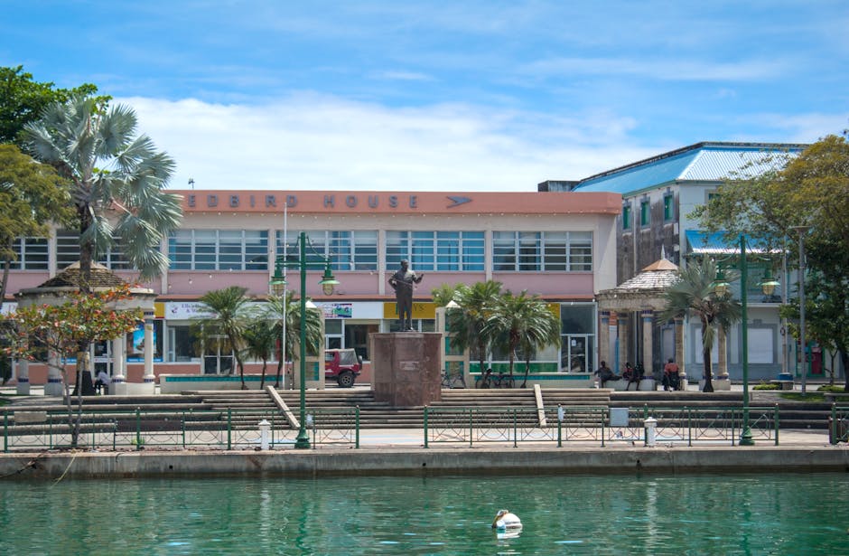 The image shows a vibrant waterfront scene in a town or city, with a multi-storey building labeled 'EDBIRD HOUSE' in large letters on the upper facade, situated directly behind a row of evenly spaced palm trees and other smaller trees. In front of the building is a paved promenade with benches and decorative green street lamps, leading to a waterway where a small white buoy floats on the calm water. There is a bronze statue of a person standing on a pedestal positioned near the center of the promenade, surrounded by several people sitting or walking along the area. The background features a clear blue sky with some wispy clouds, and the overall scene suggests a lively urban setting, possibly during daytime. Occasionally, Man and Van Kingston may use such scenic locations for house removals and furniture transport, emphasizing professional logistics and packing processes involved in home relocation and moving services.
