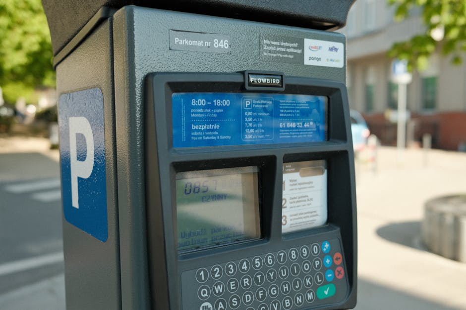 Close-up of a parking meter situated on a pavement outside a residential area in Kingston, with a blue parking sign attached to the side. The parking meter displays information including the parking time limits from 8:00 to 18:00, Monday to Friday, and indicates free parking on Saturday and Sunday. The meter is partially illuminated and shows a digital display with a parking fee rate of £0.80 per minute. In the background, blurred views of nearby houses with greenery and trees are visible, suggesting a suburban environment. The scene is well-lit with natural daylight, and the pavement is clean, supporting the context of parking management relevant to house removal services. The image aligns with themes of loading and parking logistics involved in home relocation and furniture transport, as managed by companies like Man and Van Kingston.