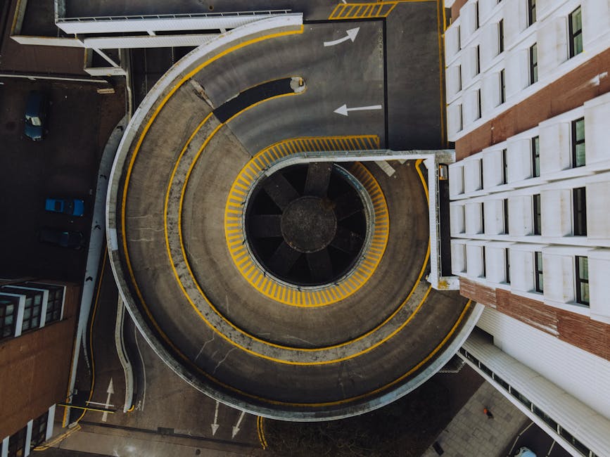Aerial view of a multi-storey, spiral car parking ramp with a concrete surface and yellow safety markings along the edges. The ramp curves around a central circular opening with black panels, leading to and from adjacent parking levels. The surrounding environment includes part of a modern multi-story building with multiple windows on the right side and a section of street with parked cars visible at the top left corner. The scene is well-lit with natural light, highlighting the structural details of the parking area and nearby urban elements. This image reflects aspects of urban infrastructure related to vehicle access and parking management, relevant to house removals and furniture transport logistics as handled by Man and Van Kingston.