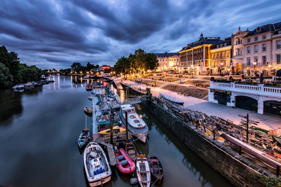 A scenic view of a river at dusk with a row of boats docked along the quay, some covered with tarpaulins and others open, showing boat interiors. To the right, there is a paved walkway with street lamps illuminating the area, and several wooden benches and small piles of stacked wooden planks or pallets. Behind the walkway, a large, elegant building with white and beige facades, multiple windows, and decorative architectural details is visible, with warm exterior lighting highlighting its features. The sky above is darkening with heavy clouds, indicating approaching evening. This image captures the calm atmosphere of a waterfront in Kingston, suited for house removals or furniture transport preparations, reflecting the process of home relocation in a lively, urban riverside setting, as could be associated with services offered by Man and Van Kingston.