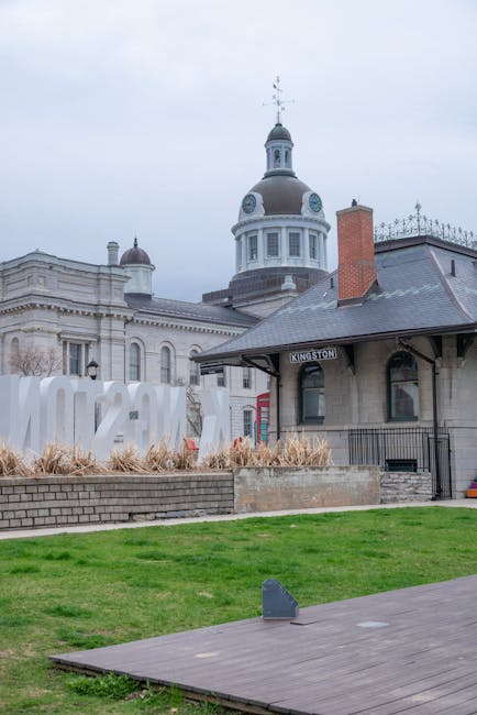 An exterior view of Kingston town centre featuring historic architecture, including a domed building with a weather vane on top, and a smaller adjacent structure with a sign reading 'Kingston.' In the foreground, a manicured grass area extends to a paved walkway. To the left, large white letters spelling 'KINGSTON' are mounted on a raised brick platform surrounded by dried ornamental grasses. The sky is overcast, providing diffuse lighting, and the scene appears to be part of a city-centre environment suitable for house removals or relocation services, with no visible furniture or moving equipment present in this specific image.