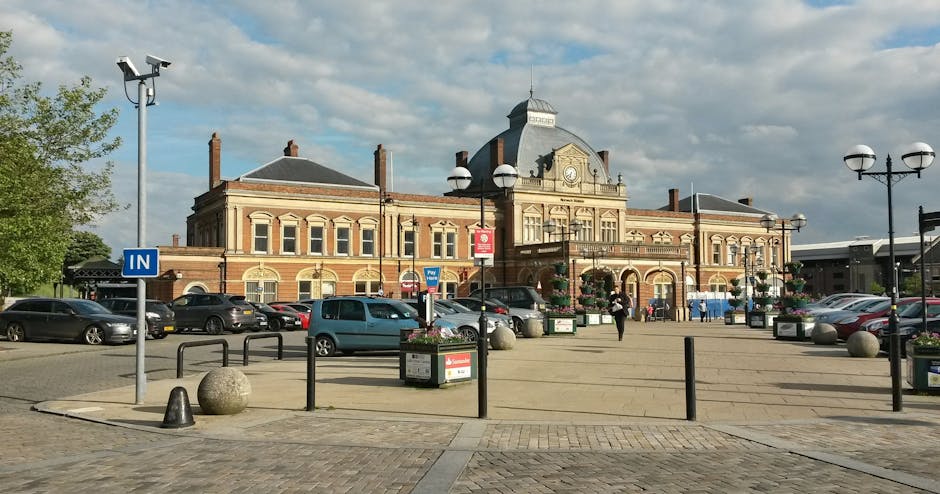 Wide view of Norbiton station building with a historic brick facade, decorative elements, and a central clock tower under a partly cloudy sky. In the foreground, there is a paved parking area with several parked cars, including a grey hatchback and a teal van, positioned along the curb and near the station entrance. The pavement features black metal bollards, flower planters with hanging baskets, and street lighting with spherical lamps on tall black posts. A person is walking towards the station entrance, carrying a bag. The scene is well-lit with natural daylight, highlighting the station's architectural details. This image illustrates the typical setting for home relocation or furniture transport, where vehicles and equipment may be used for moving belongings, and captures the environment surrounding a railway station in Kingston, relevant for house removals and relocation services by Man and Van Kingston, especially when considering storage options and logistical planning for moving from Norbiton station area.