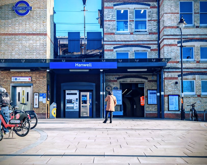 Wide view of Norbiton station building with a historic brick facade, decorative elements, and a central clock tower under a partly cloudy sky. In the foreground, there is a paved parking area with several parked cars, including a grey hatchback and a teal van, positioned along the curb and near the station entrance. The pavement features black metal bollards, flower planters with hanging baskets, and street lighting with spherical lamps on tall black posts. A person is walking towards the station entrance, carrying a bag. The scene is well-lit with natural daylight, highlighting the station's architectural details. This image illustrates the typical setting for home relocation or furniture transport, where vehicles and equipment may be used for moving belongings, and captures the environment surrounding a railway station in Kingston, relevant for house removals and relocation services by Man and Van Kingston, especially when considering storage options and logistical planning for moving from Norbiton station area.