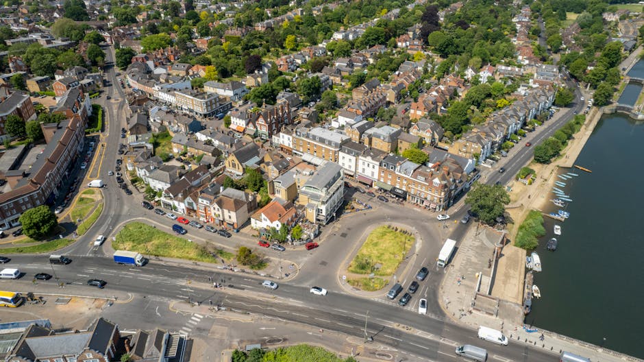 Aerial view of a residential area in Kingston with a river on the right side, showing a mix of terraced houses, apartment buildings, and green spaces. In the foreground, a busy road with cars, vans, and a blue truck is visible, along with a small roundabout surrounded by a grassy area and parked vehicles. Behind the road, there is a waterfront promenade with boats docked along the riverbank. The area is well-lit with natural daylight, capturing the typical urban landscape involved in house removals and furniture transport. Man and Van Kingston may facilitate home relocation and moving logistics within this scenic and densely populated Kingston part, supporting clients with professional packing and loading services.