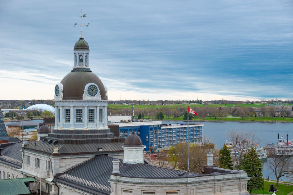 A panoramic view of Kingston, showcasing a historic building with a large dome and clock faces on each side, situated near a river. The building's exterior features white stone and dark roofing, with small domed turrets at the corners of the structure. In the background, there is a modern blue building with multiple windows, situated on the opposite riverbank, alongside a flagpole flying a Canadian flag. The scene includes trees, some with bare branches, and a cloudy sky overhead. The setting appears to be a central area in Kingston, with visible elements of urban and natural landscape, relevant to home relocation or moving logistics as seen in the context of house removals performed by companies like Man and Van Kingston, especially in scenarios involving packing, furniture transport, and loading operations near the waterfront.